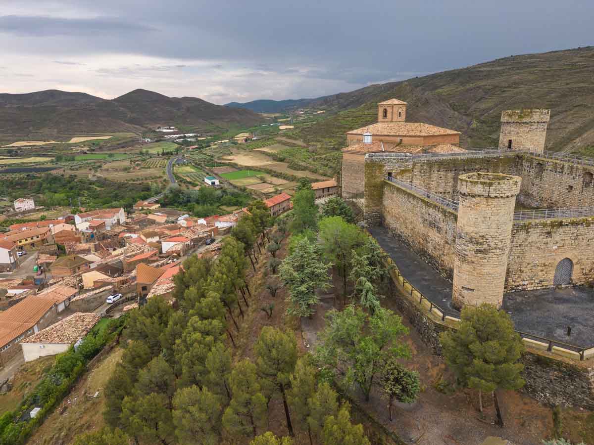 Vista aérea del Castillo de Cornago y la Iglesia de San Pedro en la Comarca del Alhama-Linares, con el pueblo a la izquierda y un paisaje de colinas al fondo.