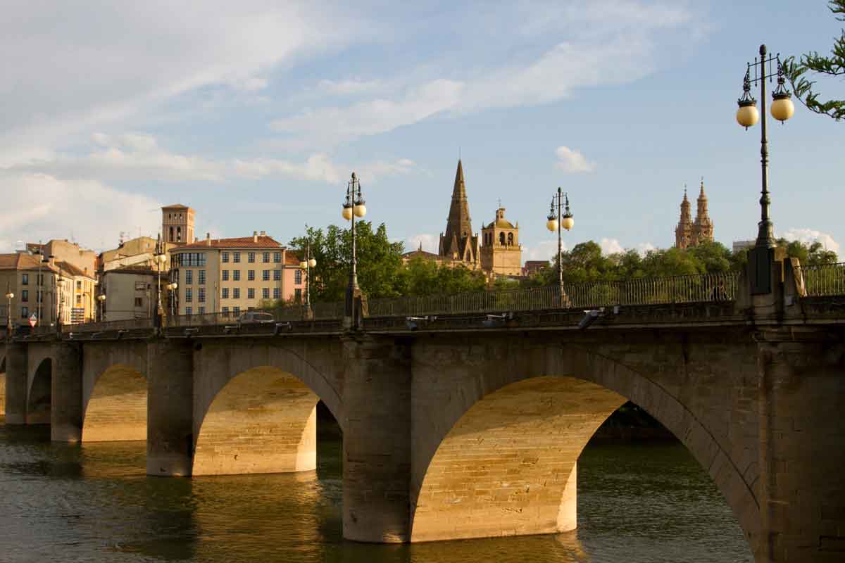 Vista del Puente de Piedra sobre el río Ebro en Logroño al atardecer, con las torres de la Concatedral de la Redonda y la aguja de la iglesia de Palacio al fondo.