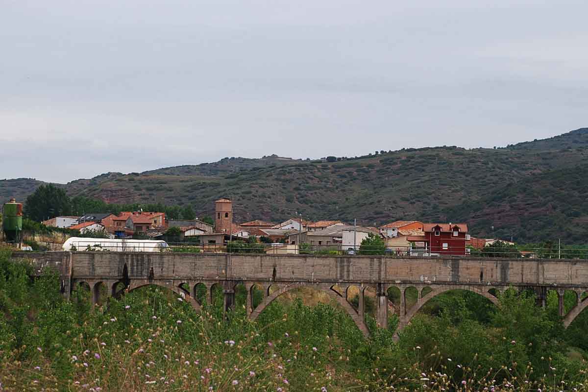 Vista de Bobadilla tras un puente de arcos, con la torre de la iglesia y las laderas del valle del Najerilla al fondo.