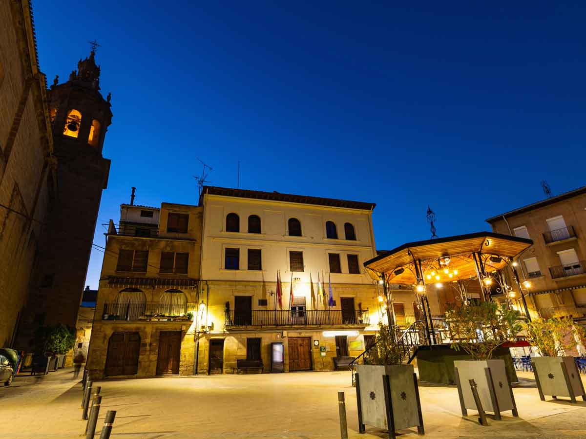 Plaza principal de Cenicero en La Rioja iluminada de noche, con el ayuntamiento, el kiosco de música y la torre de la iglesia bajo un cielo de hora azul.
